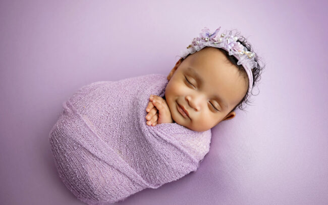 Sleeping newborn baby wrapped in lavender with a floral headband on a purple backdrop