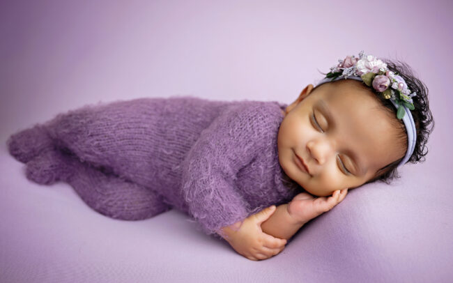 Newborn baby posed on their side in a soft purple outfit and floral headband