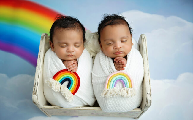 Newborn twins wrapped in white and posed together in a small bed with a rainbow backdrop