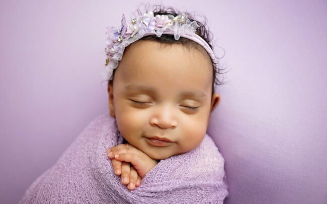 Close-up portrait of a sleeping newborn baby in a lavender wrap and jeweled floral headband