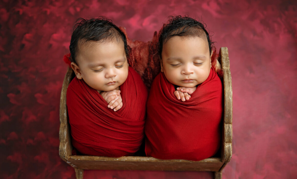 Sleeping newborn twins wrapped in red and posed together in a wooden newborn bed