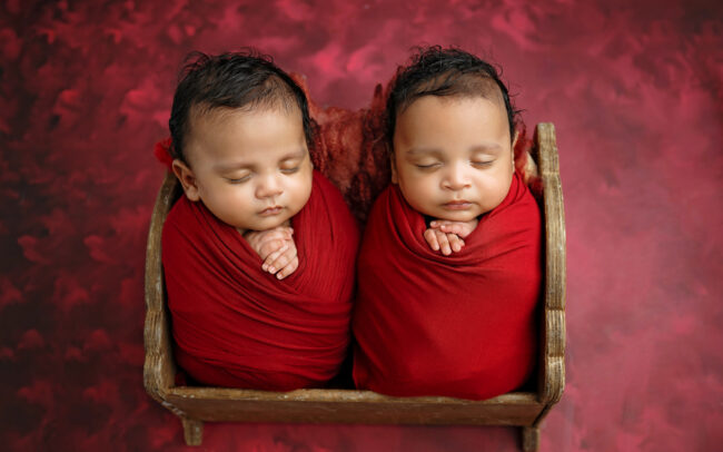 Sleeping newborn twins wrapped in red and posed together in a wooden newborn bed