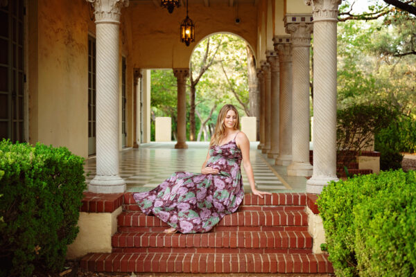 Pregnant mother in a floral dress sitting on brick steps beneath the arched columns at Landa Library in San Antonio.