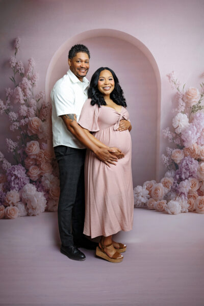 Expecting couple posing together on a soft pink floral backdrop during their San Antonio maternity photography session.