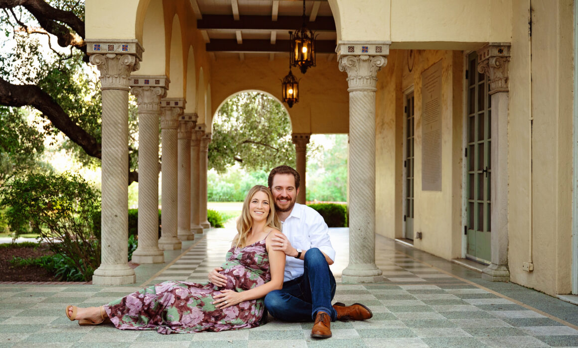 Expecting couple seated beneath the columns of Landa Library during a romantic San Antonio maternity photography session.