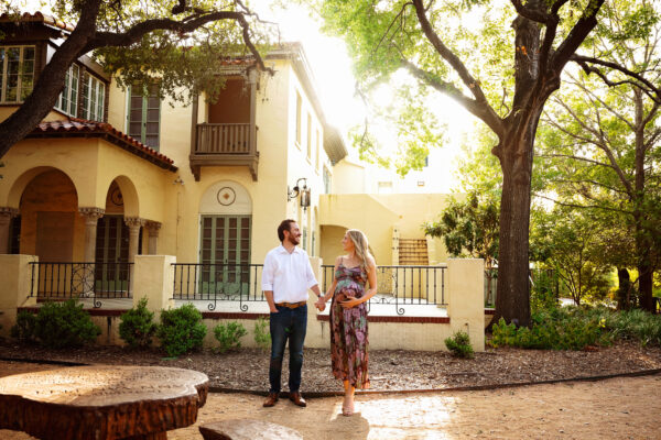 Expecting parents walking hand in hand beneath large oak trees during a sunset maternity photoshoot in San Antonio.