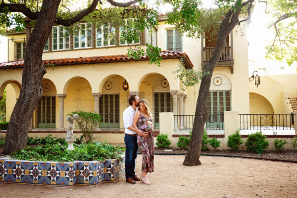 Father kissing expectant mother beside the Spanish-style architecture and courtyard gardens at Landa Library.