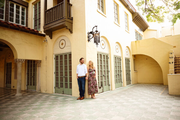 Pregnant woman and partner standing beside the arched windows and historic architecture of Landa Library in San Antonio.