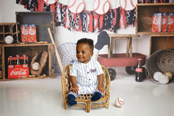 Baby sitting in a small wicker chair surrounded by vintage baseball props for an 8-month milestone portrait