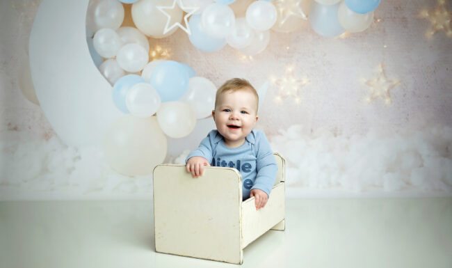 Six month old baby smiling in a vintage white bed prop with soft blue moon and balloon backdrop.