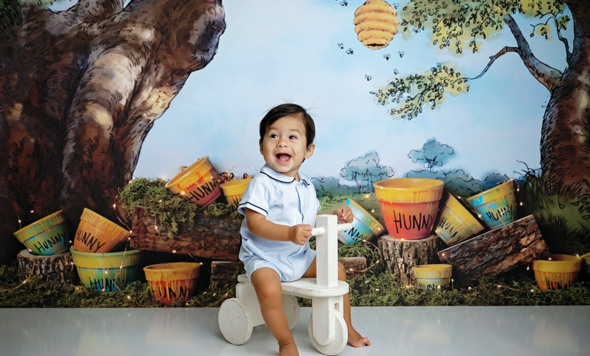 Baby boy smiling on a small white tricycle with a Winnie the Pooh-inspired first birthday backdrop.
