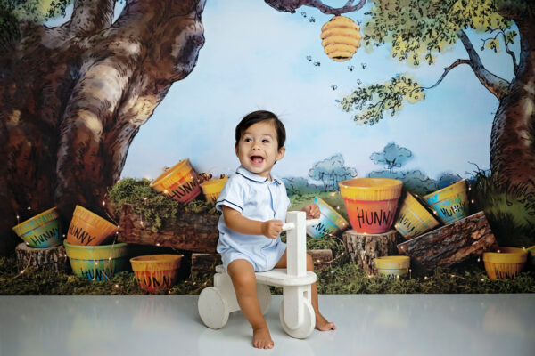 Baby boy smiling on a small white tricycle with a Winnie the Pooh-inspired first birthday backdrop.
