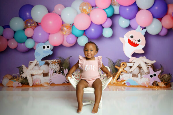 One-year-old girl smiling and standing in front of a colorful Baby Shark birthday backdrop.