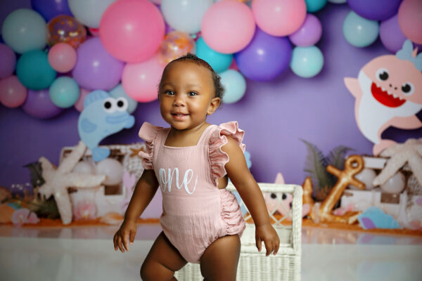 Smiling one-year-old girl standing during her pink and purple Baby Shark first birthday session.