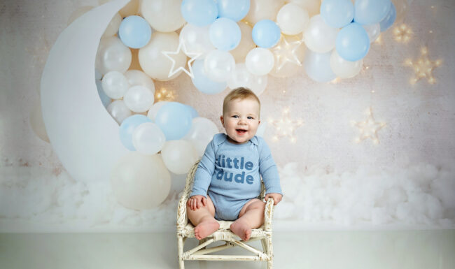Six month old baby sitting in a white wicker chair with blue balloons, moon, stars, and cloud backdrop.
