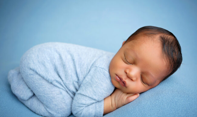 Newborn baby sleeping peacefully in a light blue romper with hands tucked beneath the face.