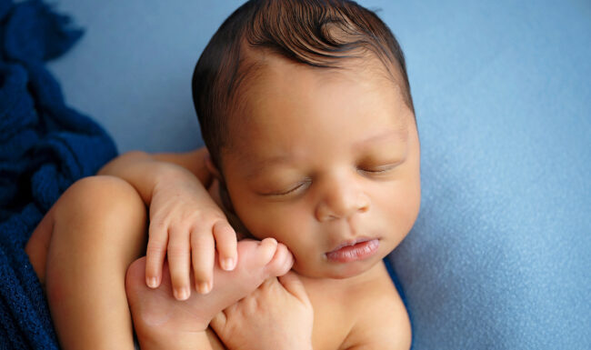 Sleeping newborn posed in a light blue romper while gently holding tiny feet on a blue backdrop.