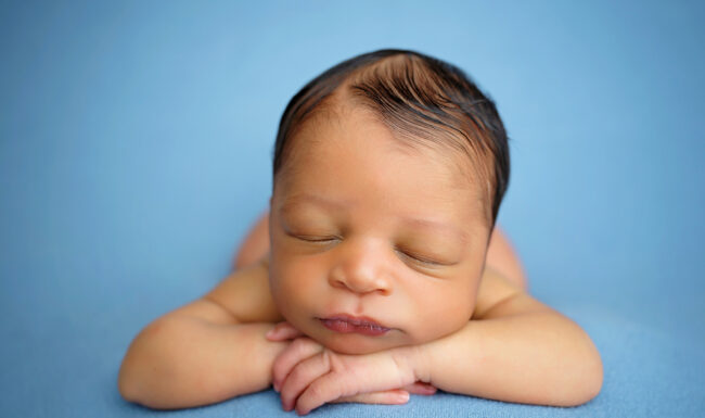 Newborn baby resting with chin above folded hands against a simple soft blue backdrop.