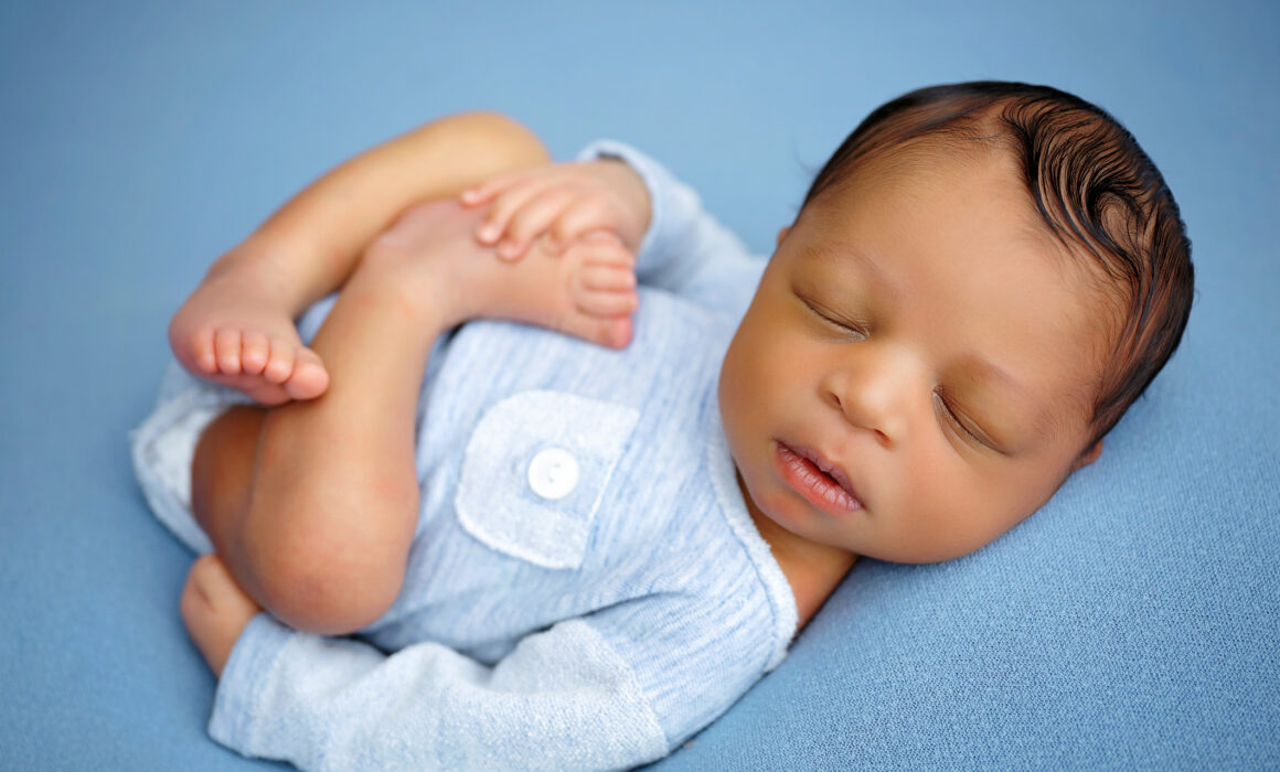 Sleeping newborn posed in a light blue romper while gently holding tiny feet on a blue backdrop.