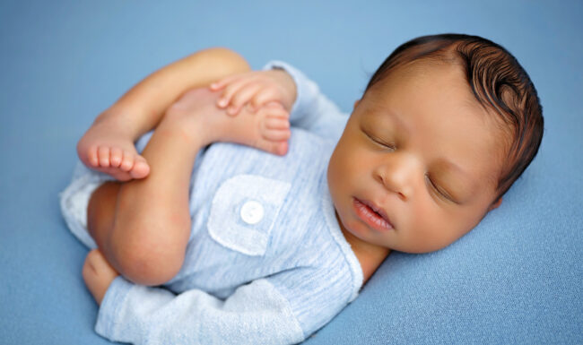 Sleeping newborn posed in a light blue romper while gently holding tiny feet on a blue backdrop.