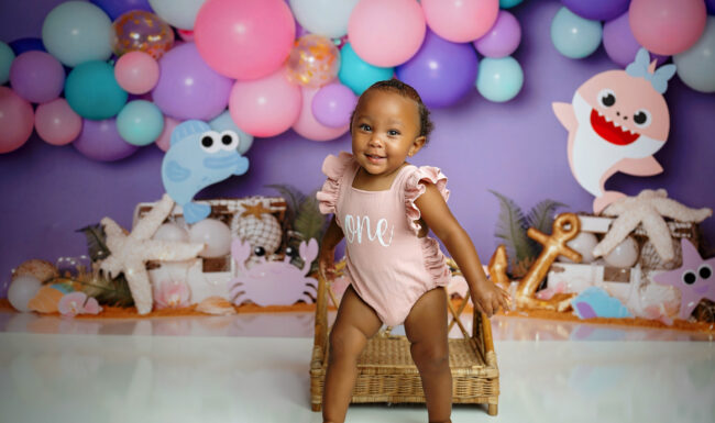 One-year-old girl smiling and standing in front of a colorful Baby Shark birthday backdrop.