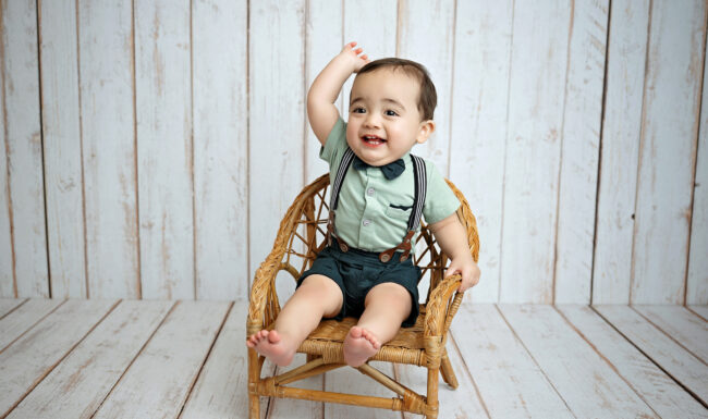 One-year-old boy smiling in a wicker chair during simple studio milestone portraits