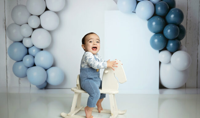 Baby boy posing on a white rocking horse with blue balloon decor for his first birthday session
