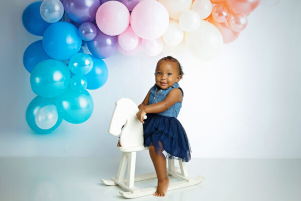 ne-year-old girl standing beside a white rocking horse with pastel balloon decorations during her milestone session.