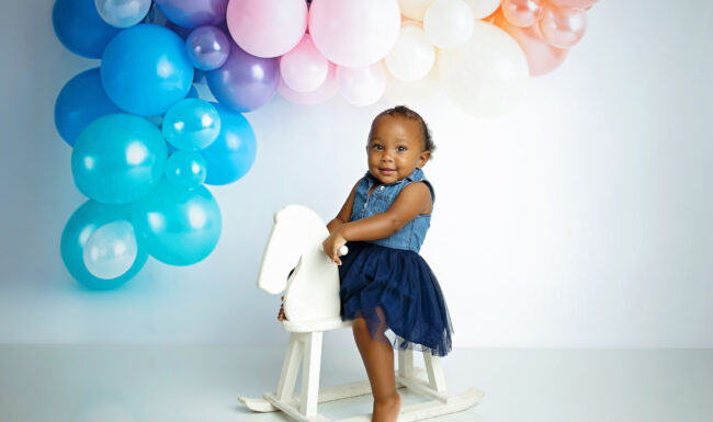 ne-year-old girl standing beside a white rocking horse with pastel balloon decorations during her milestone session.