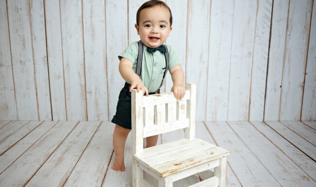 Smiling baby boy standing behind a white chair during his first birthday milestone photography session