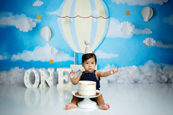 Baby boy celebrating his first birthday with a white cake and hot air balloon cake smash backdrop.