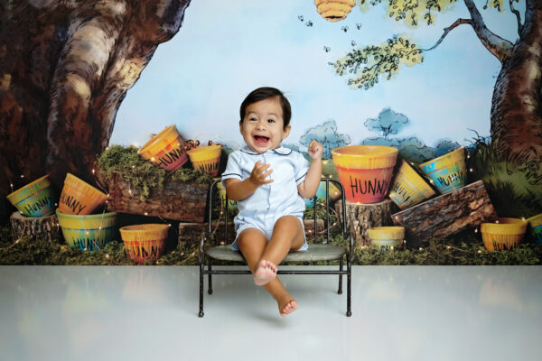 Happy one-year-old boy sitting in front of a Winnie the Pooh-inspired first birthday photography setup.