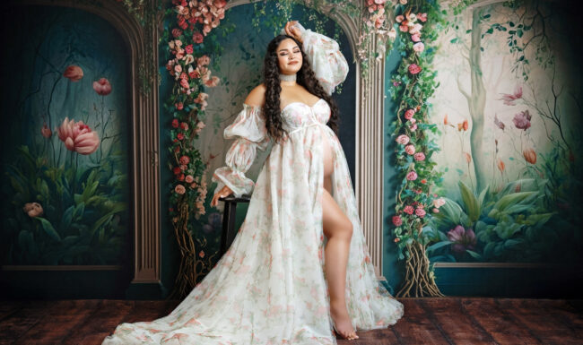 Expecting mother posing in a floral maternity gown with a romantic garden backdrop in a San Antonio studio