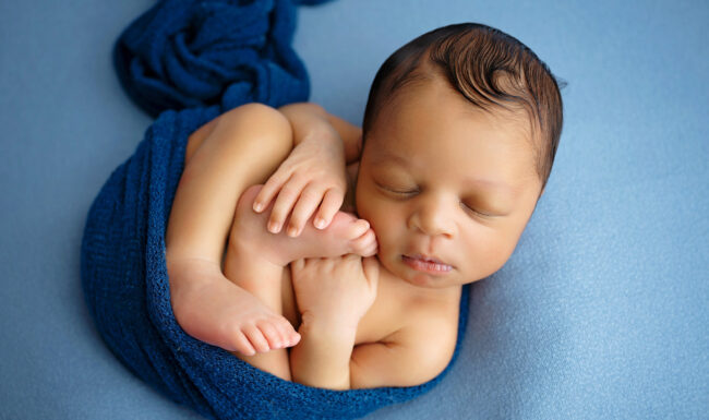 Newborn baby curled on a soft blue backdrop with a deep blue wrap and tiny hands and feet visible.