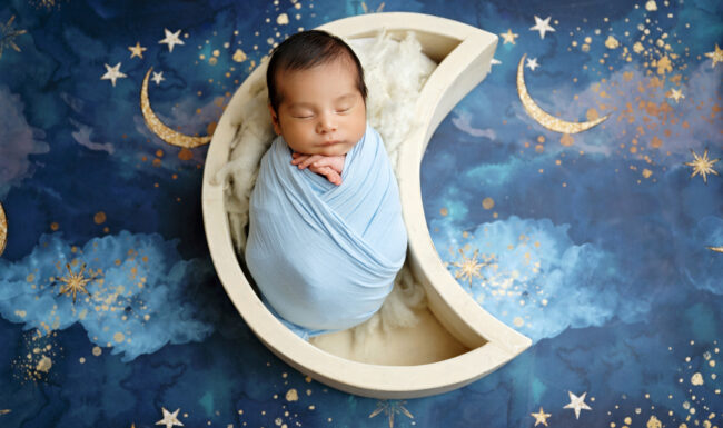 Newborn baby wrapped in light blue in a crescent moon prop surrounded by blue stars and celestial details.