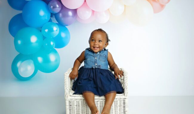 Smiling one-year-old girl in a blue dress sitting on a white chair beneath pastel birthday balloons.
