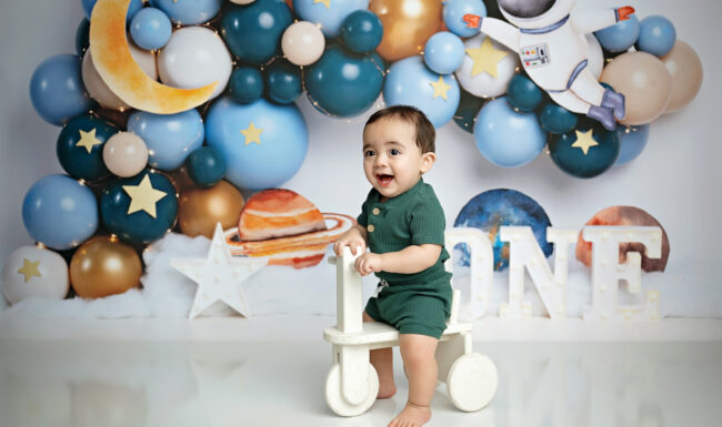 Baby boy posing with a ride-on toy in front of an astronaut and space themed first birthday backdrop