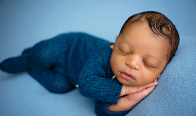 Newborn baby sleeping peacefully in a light blue romper with hands tucked beneath the face.