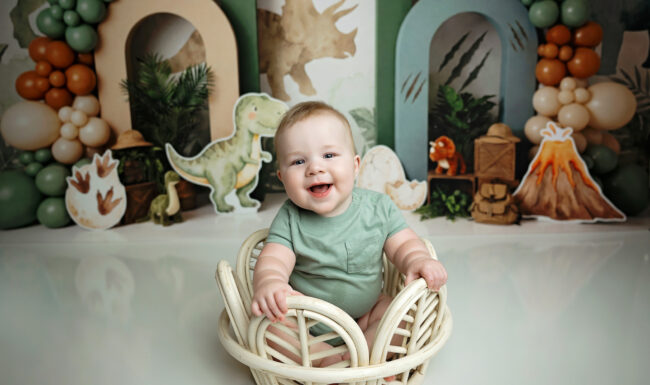 Smiling six month old baby sitting in a white wicker basket during a dinosaur milestone photography session in San Antonio.