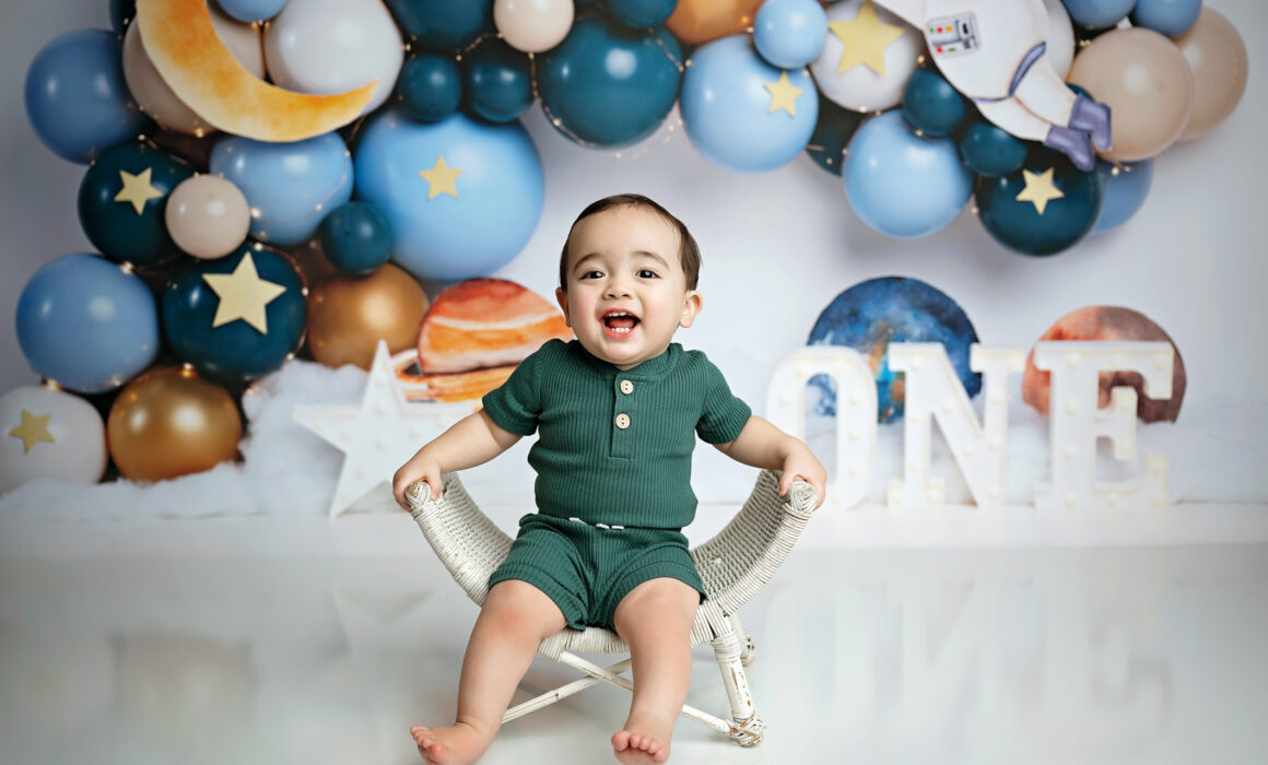Smiling one-year-old boy in front of a blue and gold outer space cake smash backdrop with ONE letters