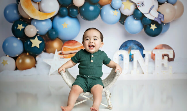 Smiling one-year-old boy in front of a blue and gold outer space cake smash backdrop with ONE letters