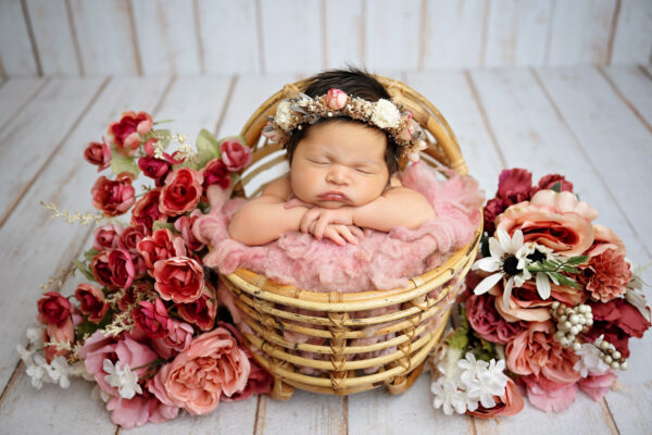 Baby girl sleeping in wicker basket surrounded by pink and burgundy flowers during San Antonio newborn photoshoot