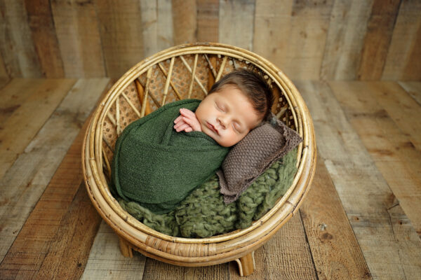 Sleeping baby boy wrapped in green on a round rattan chair during a newborn photography session.
