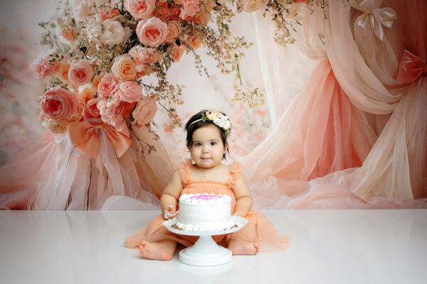Baby girl wearing a peach dress seated behind a white birthday cake surrounded by blush pink flowers during her San Antonio cake smash session.