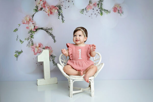 Baby girl sitting in a white chair during her one-year milestone portraits in a San Antonio studio