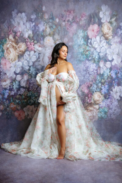 Expecting mother wearing a flowing floral maternity gown against a romantic floral backdrop