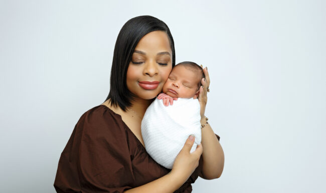 Mother gently holding her swaddled newborn baby boy against her cheek in a studio portrait.