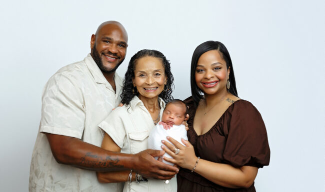 Proud family members gathered around a sleeping newborn baby during a multigenerational studio portrait.