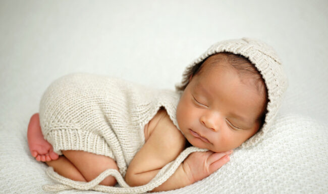 sleeping newborn in a cream knit romper and bonnet posed on a soft ivory textured blanket.