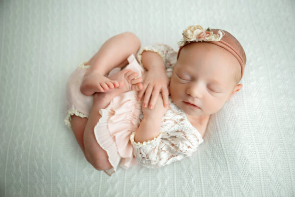 Newborn baby girl curled up in a delicate cream lace outfit with a blush pink floral headband on a soft white blanket.
 